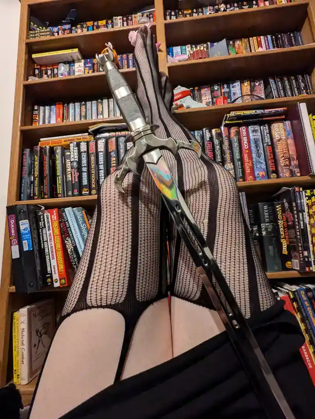 A person wearing black fishnet stockings and a black top is seated in front of a wooden bookshelf filled with fantasy and…