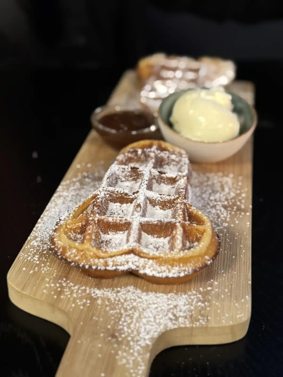 A golden-brown waffle shaped like a Christmas tree sits on a wooden serving board, dusted with powdered sugar. In the…