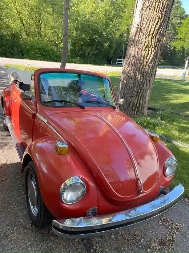 A red Volkswagen Beetle convertible is parked on a gravel driveway next to a large tree, with green grass and trees in the…