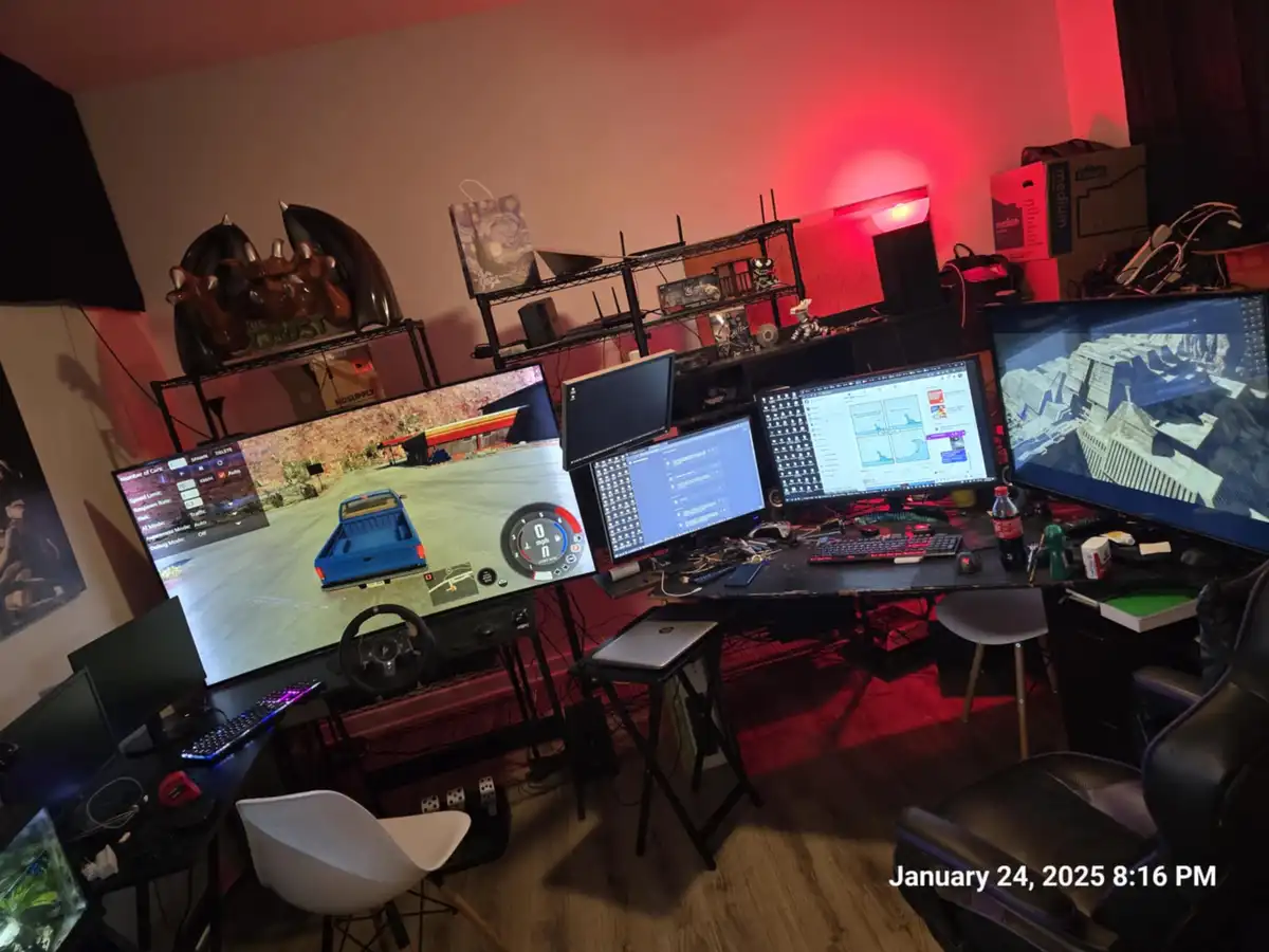 A cluttered, red-lit gaming room features a large desk with multiple monitors displaying various activities: a racing game…