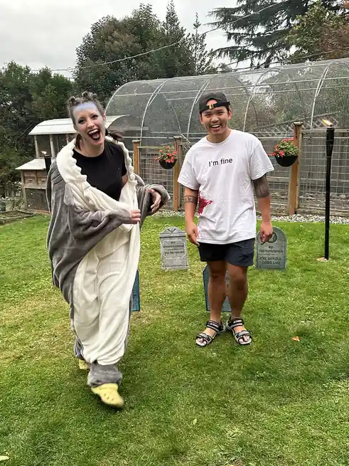 Two people stand in a grassy yard decorated with fake tombstones and torches. The person on the left wears a grey and white…