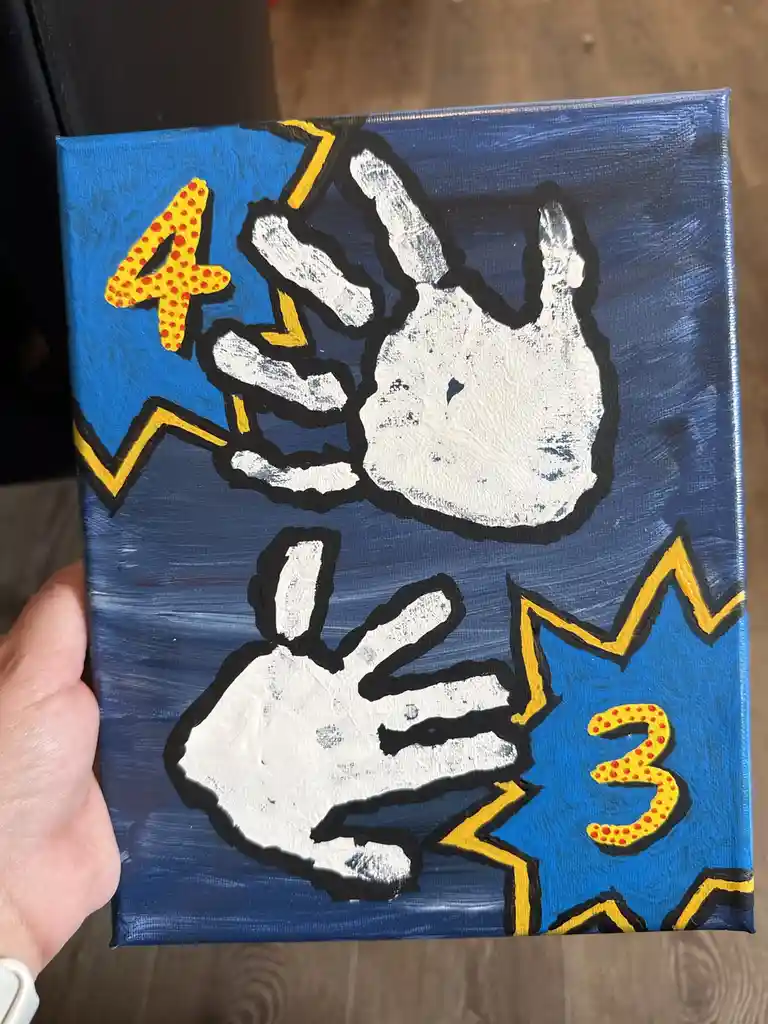 A hand-painted canvas depicts two white handprints outlined in black against a blue background, with a yellow lightning bolt…