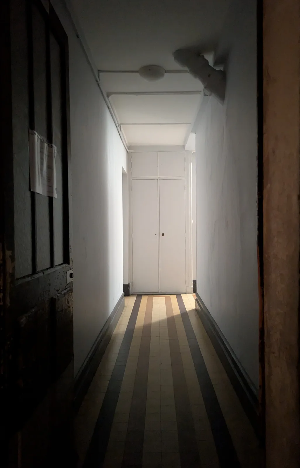 A dimly lit, narrow hallway with striped tile flooring leads to a white door at the end. The walls are plain white, and a…