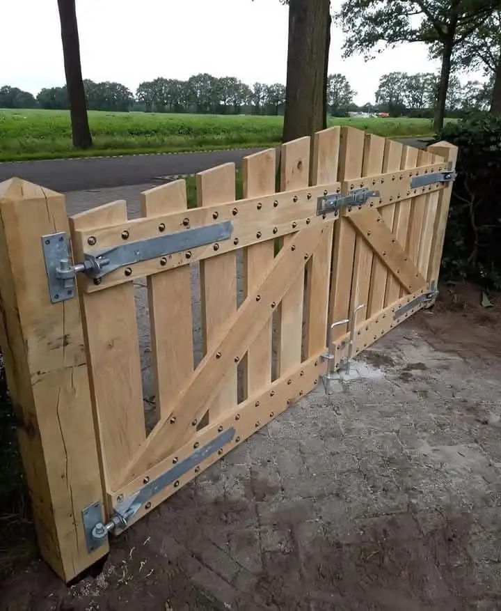a large wooden gate with metal hinges and bolts is shown leaning open on a paved path, with a grassy field and trees in the…