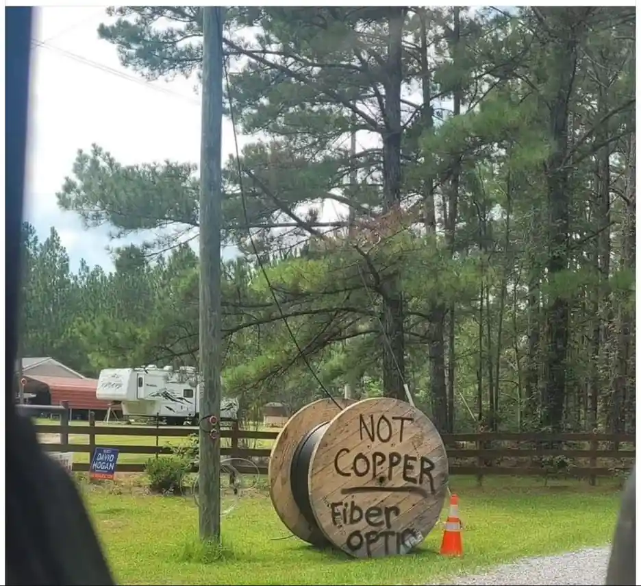 A large wooden spool lying on grass in a rural setting, with handwritten text reading 'NOT COPPER Fiber OPTIC'. Behind it…