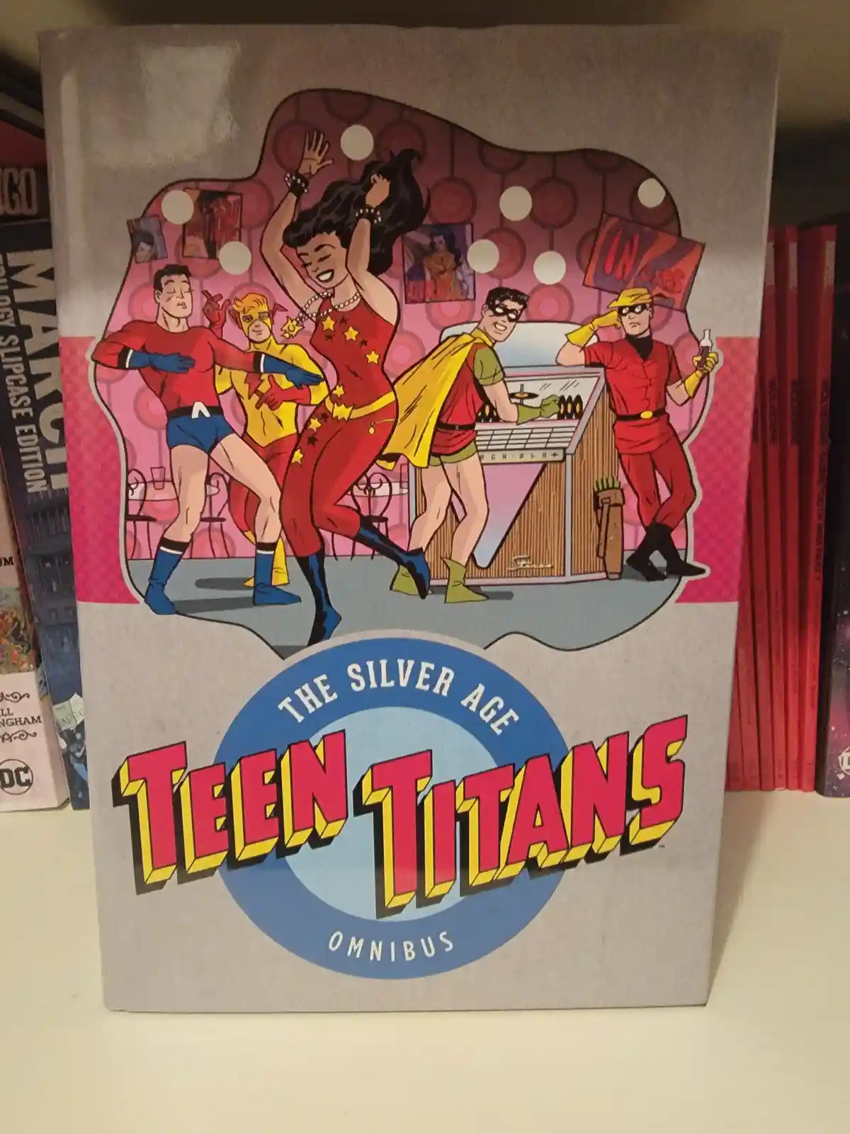 The cover of 'The Silver Age Teen Titans Omnibus' comic book shows five superheroes in 1960s-style costumes dancing and…