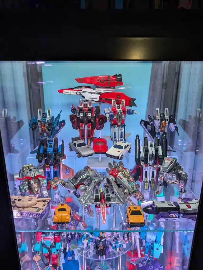 A glass display case filled with numerous Transformers action figures and vehicles, arranged on multiple shelves. The…