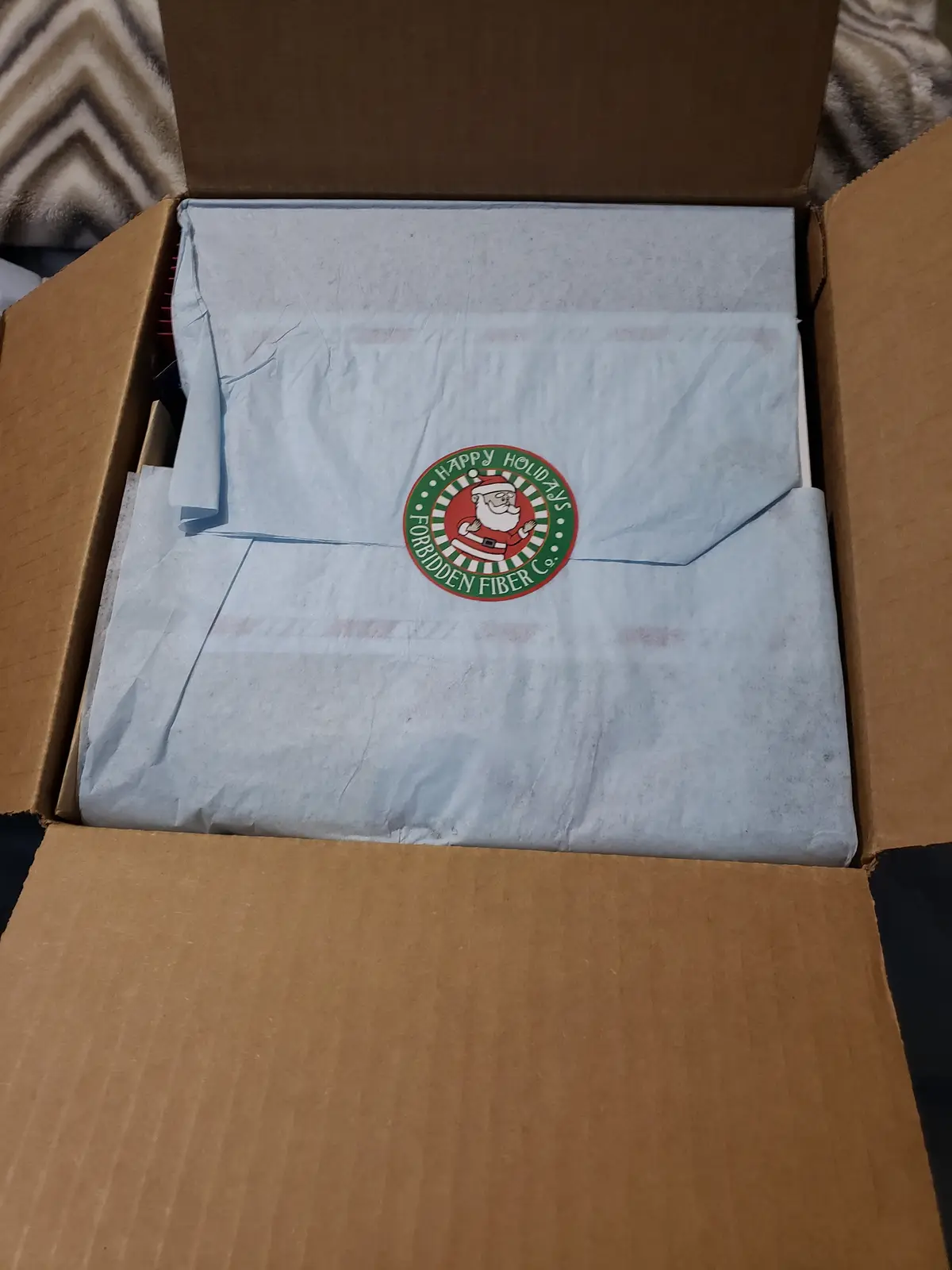 An open cardboard box contains a package wrapped in light blue tissue paper, which has a circular holiday sticker. The…
