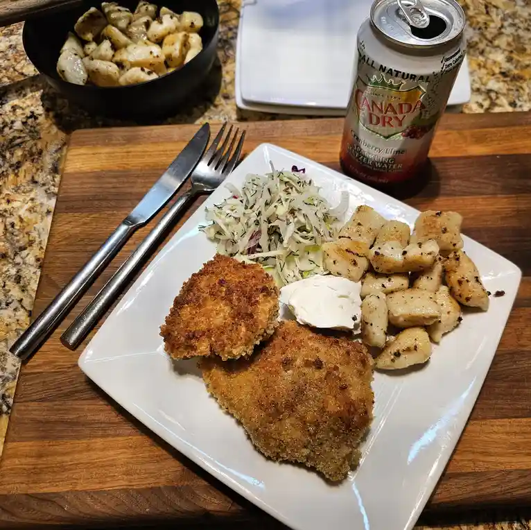 A plated meal featuring two breaded and fried cutlets, a side of gnocchi, a small portion of coleslaw, and a dollop of sour…