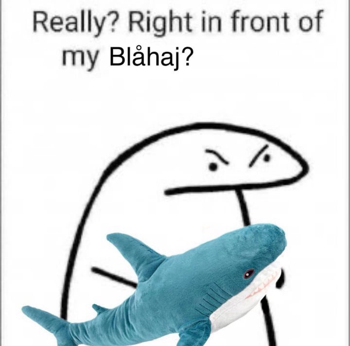 A simple black-and-white stick figure with an angry, skeptical expression stares down at a blue plush shark toy. The plush…