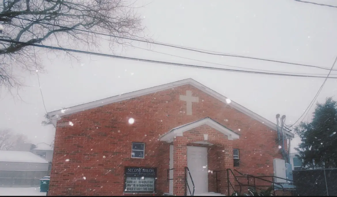 A snowy day outside a red brick church with a white cross on its gable, identified by a sign as 'SECOND SHILOH MISSIONARY…