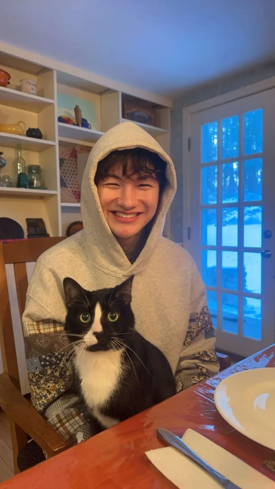 A young person in a light gray hoodie sits at a dining table, winking and smiling broadly while holding a black-and-white…