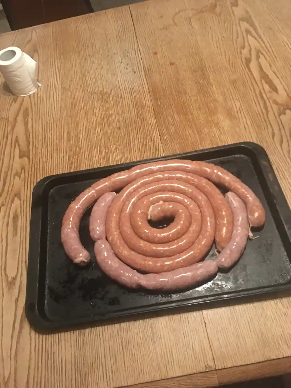 A black baking tray holds a large spiral of raw sausages on a wooden table, with a roll of toilet paper visible in the…