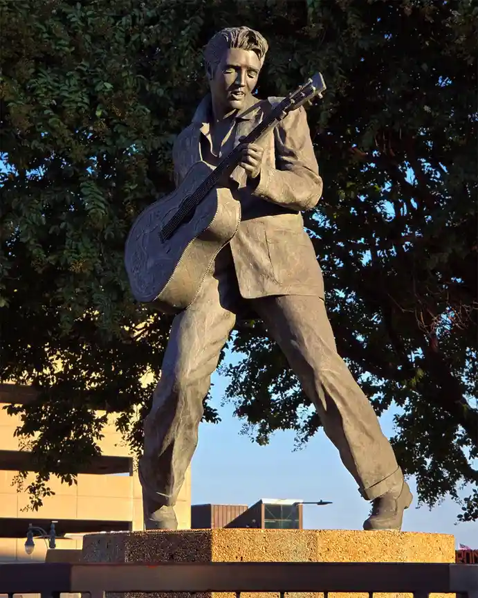 A bronze statue of Elvis Presley stands on a pedestal, posed mid-performance while holding an acoustic guitar with 'ELVIS'…