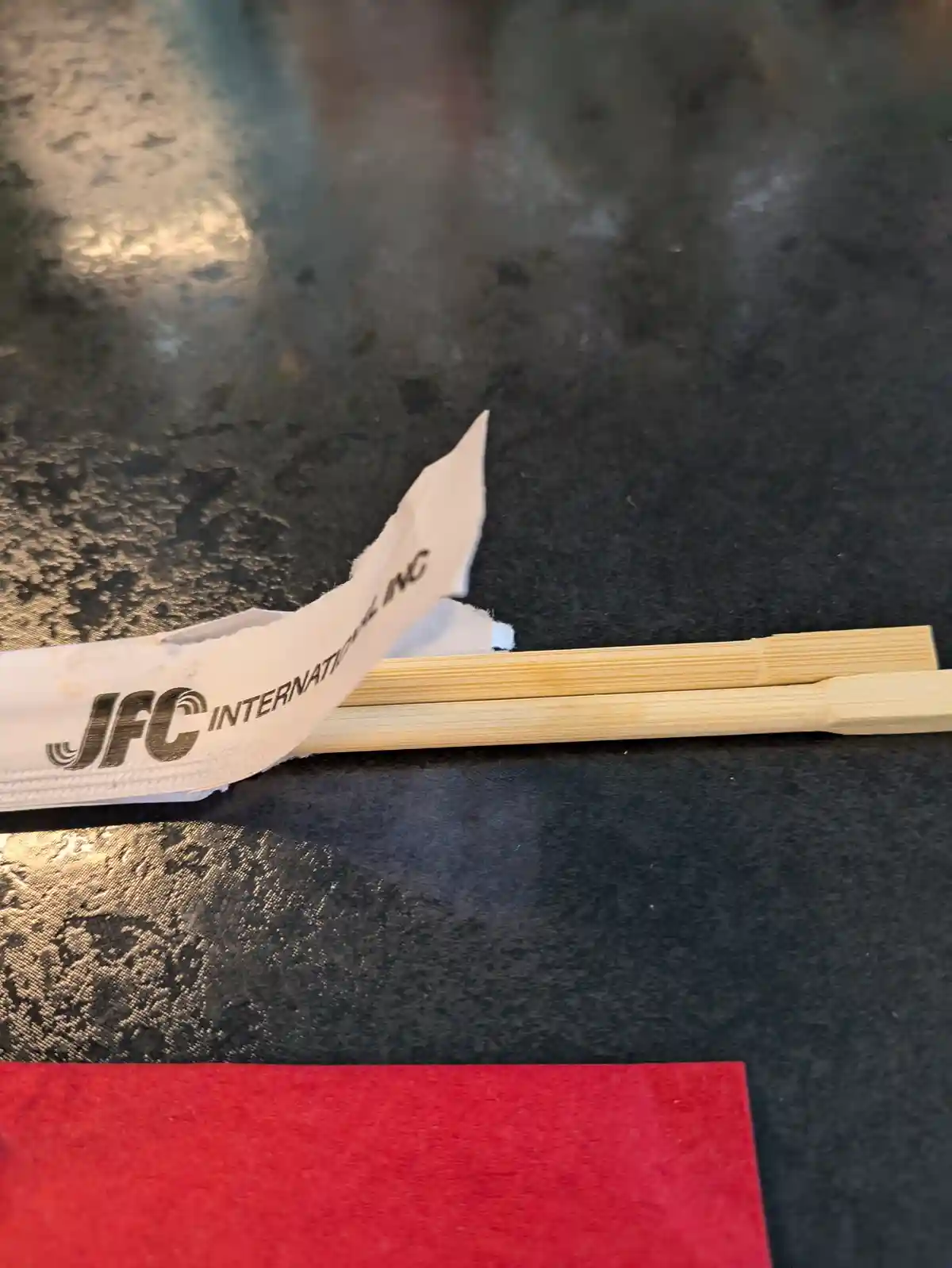 A close-up photo shows a pair of light-colored bamboo chopsticks resting on a dark, textured surface next to a torn white…