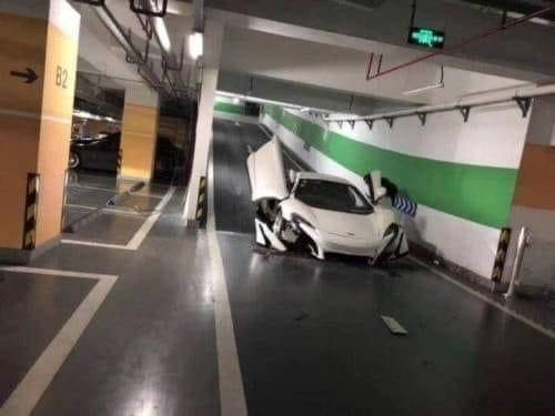 A white sports car is severely damaged after crashing into a wall in an underground parking garage, with its front end…