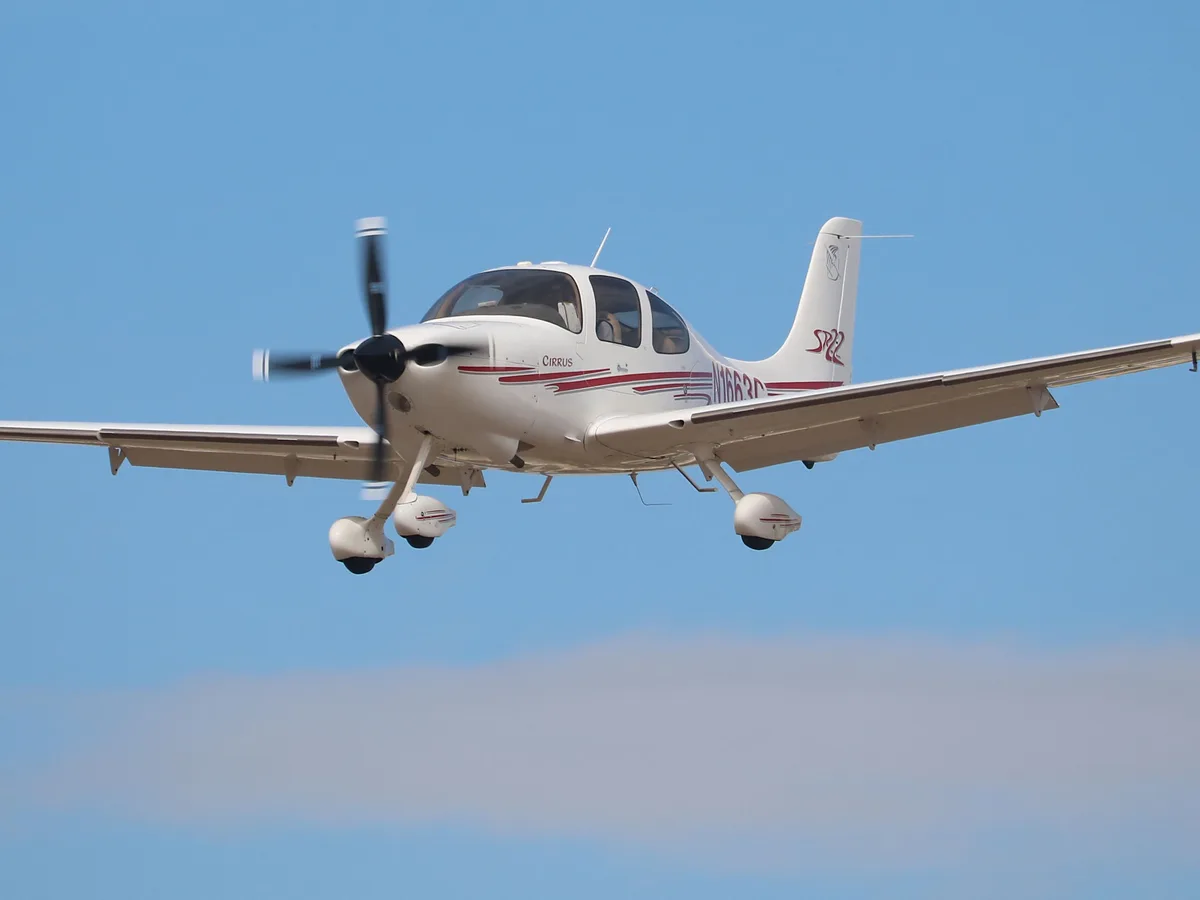 A white Cirrus SR22 single-engine propeller airplane is captured in flight against a clear blue sky with a thin cloud layer…