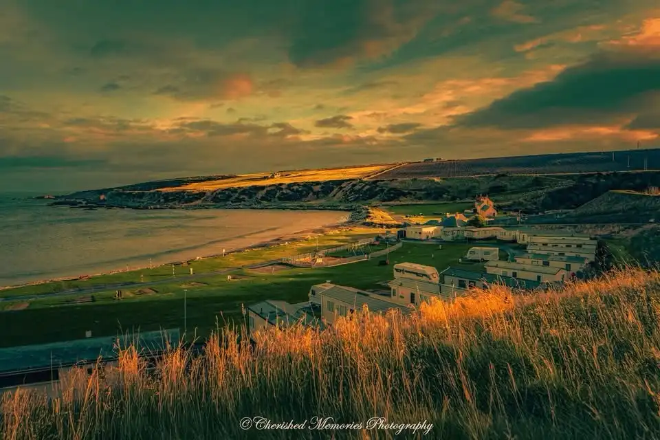 A scenic coastal landscape at sunset with golden light illuminating grass, cliffs, and a caravan park near the water…