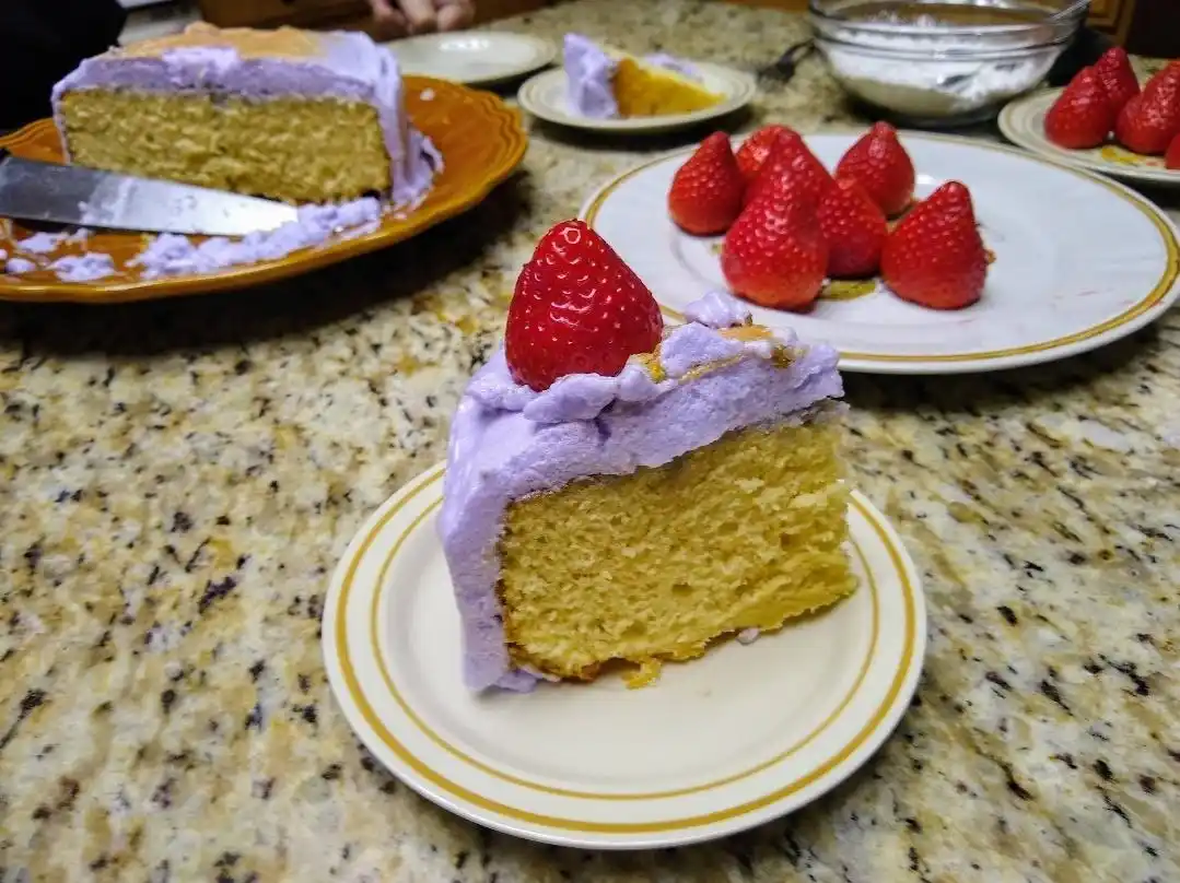 A slice of yellow cake with purple frosting and a single strawberry on top sits on a white plate with gold trim, placed on a…