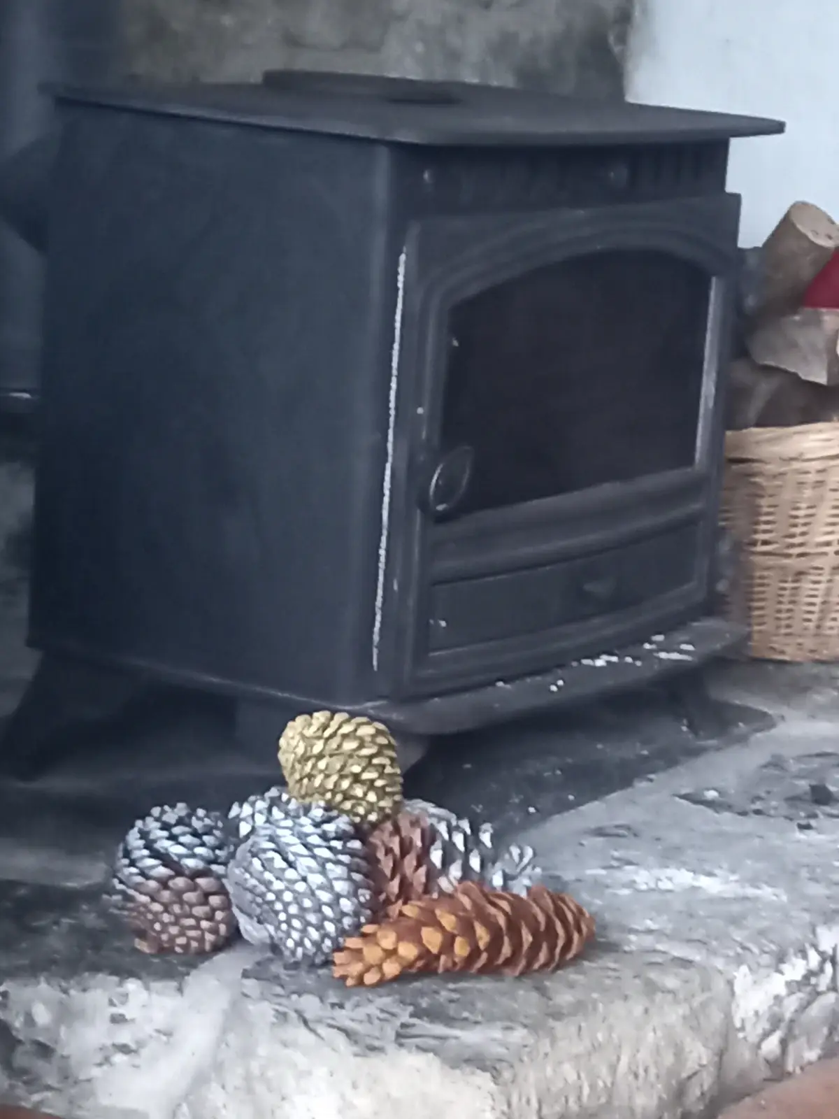 A black metal wood-burning stove sits on a stone hearth, with several pinecones of varying sizes and colors—some natural…