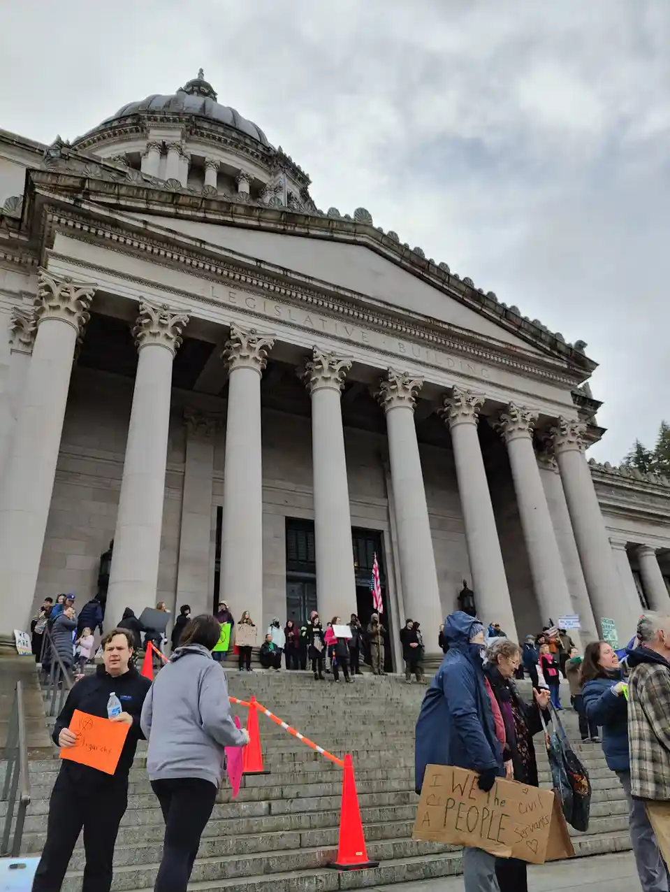A protest scene outside a large neoclassical building labeled 'LEGISLATIVE BUILDING' with many people holding signs. One…