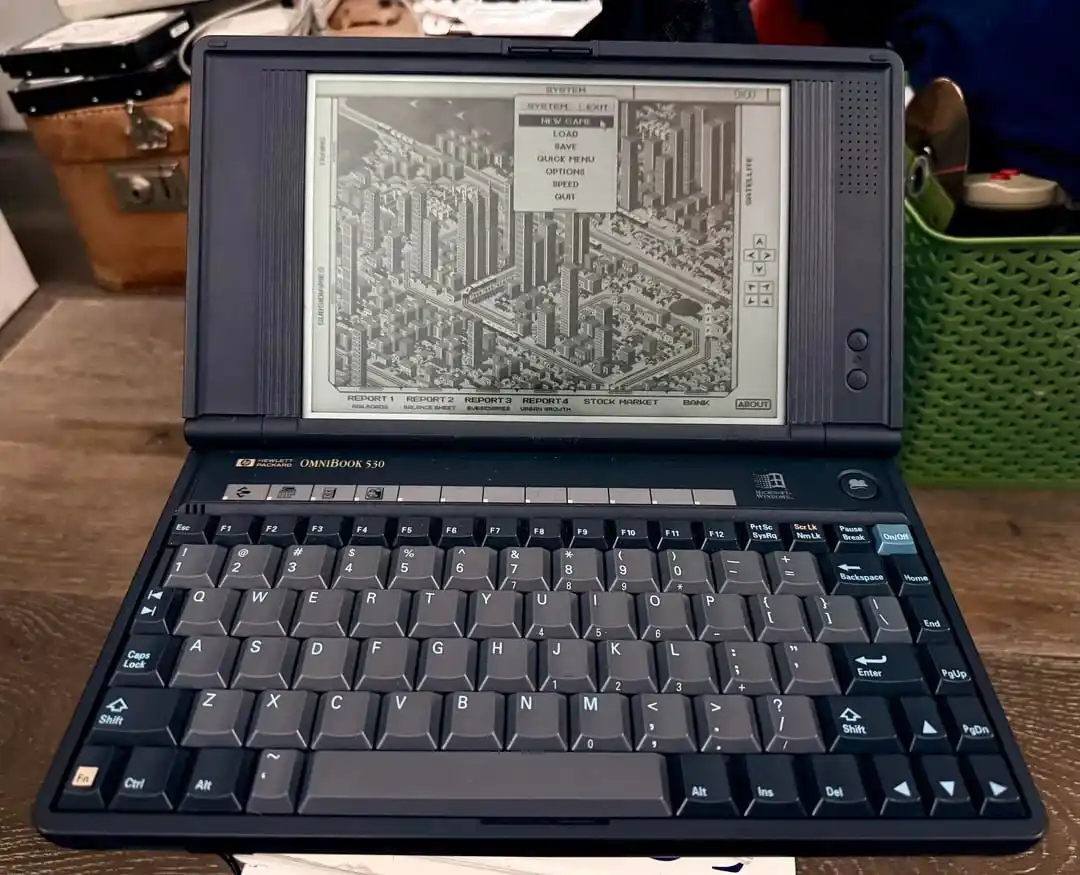 A black Hewlett-Packard OmniBook 530 laptop displays a grayscale cityscape game interface with a menu overlay listing…