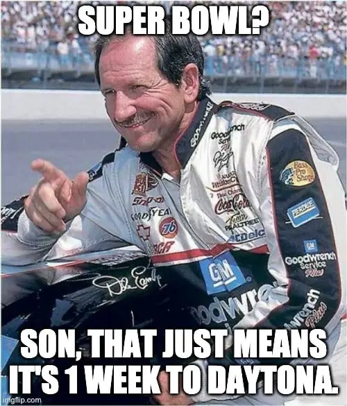A man in a NASCAR driver's racing suit, leaning on his car with a smug, knowing smile, points at the viewer. The image has…