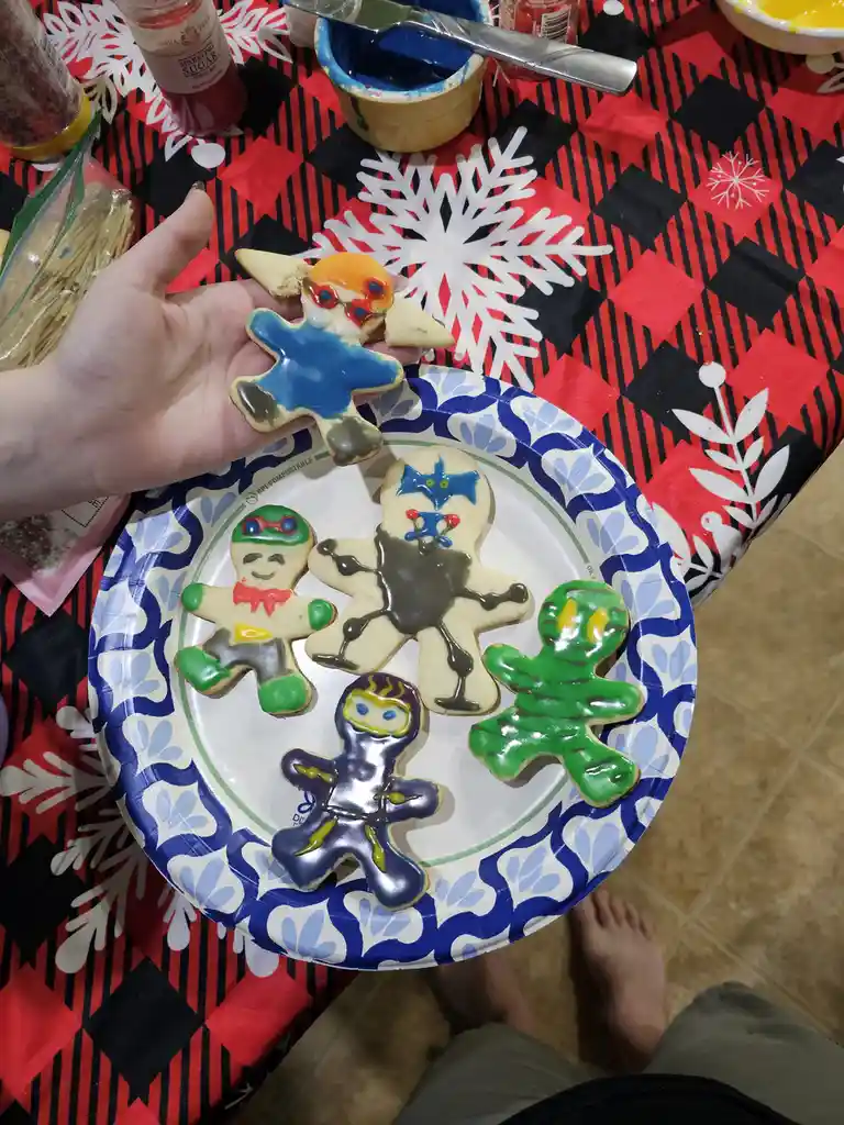A hand holds up a badly decorated gingerbread cookie with melted icing, while a paper plate below displays four other…