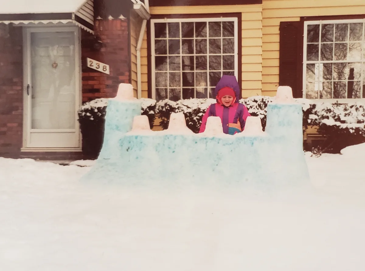 A child in a colorful winter coat and hat stands behind a large, snow-covered castle structure in front of a house. The…