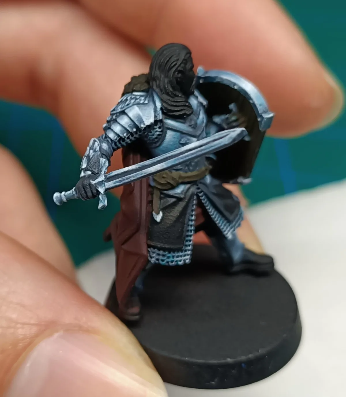 A close-up photo shows a hand holding a painted miniature figurine of a medieval-style warrior in silver armor, holding a…