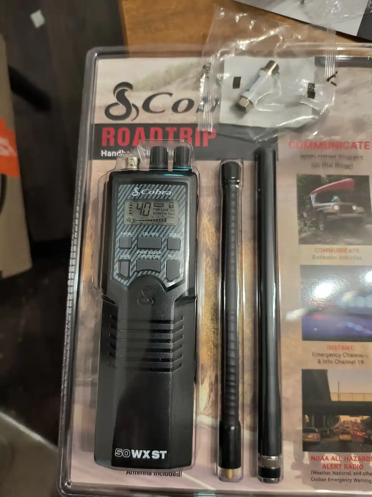 A Cobra Roadtrip handheld CB radio, model 50WX ST, is shown in its original plastic packaging with two antennas and…