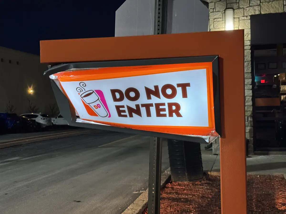 A brightly lit orange and white 'DO NOT ENTER' sign, featuring a coffee cup logo with 'DD' initials, is visibly bent and…