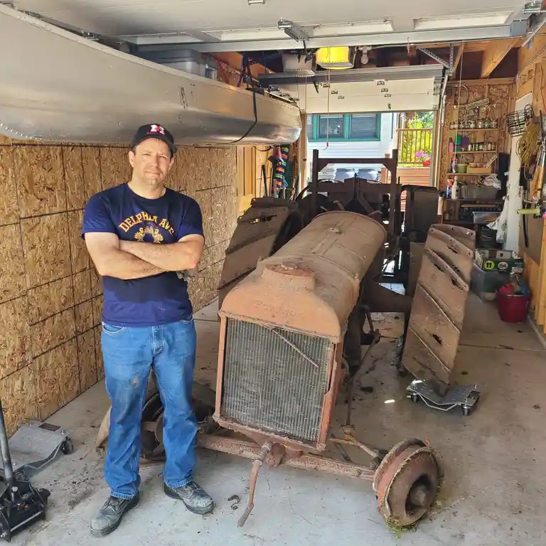 A man in a blue t-shirt and jeans stands with arms crossed in a cluttered garage next to a rusty, disassembled vintage…