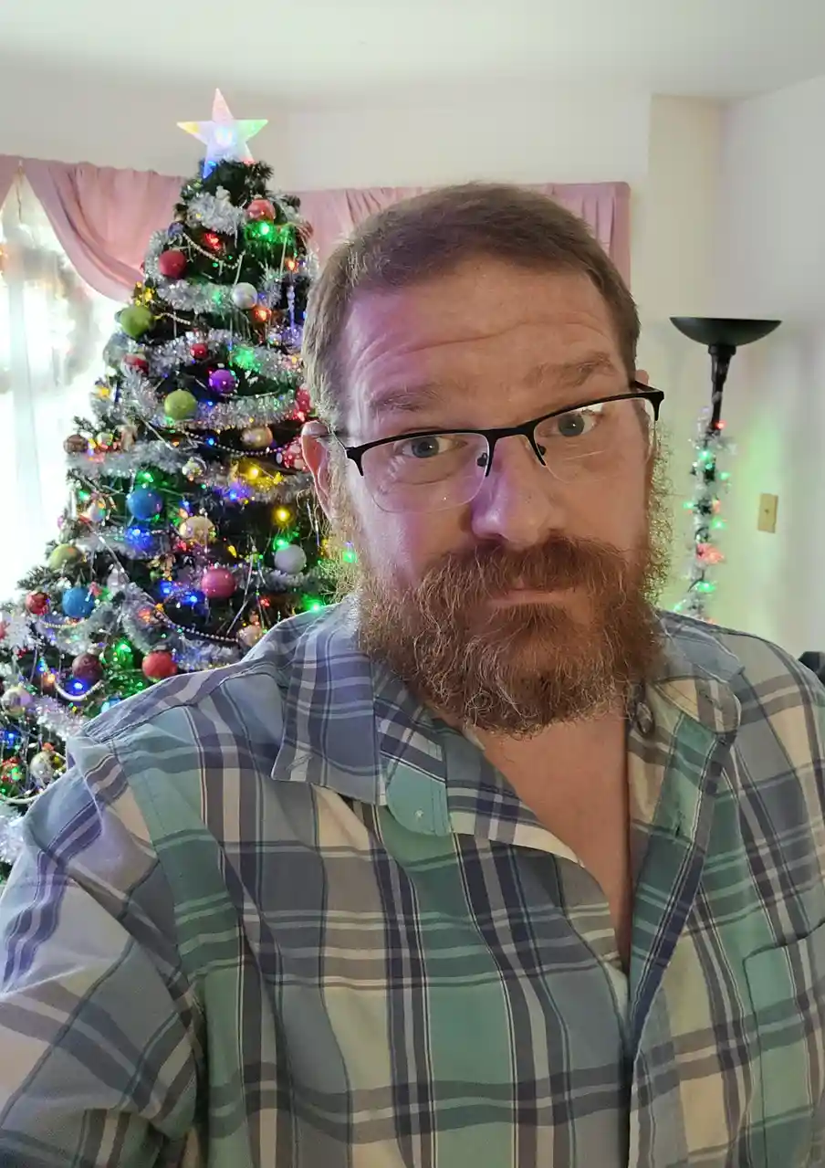 A man with a beard and glasses stares directly at the camera with a skeptical, unimpressed expression. Behind him is a…