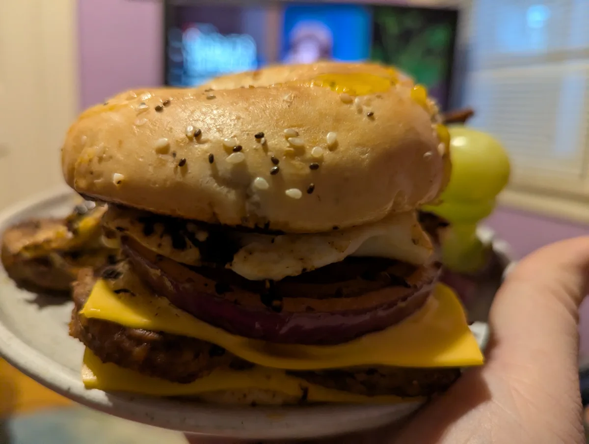 A close-up photo shows a hand holding a plate with a towering bagel burger. The bagel, topped with sesame and chia seeds, is…