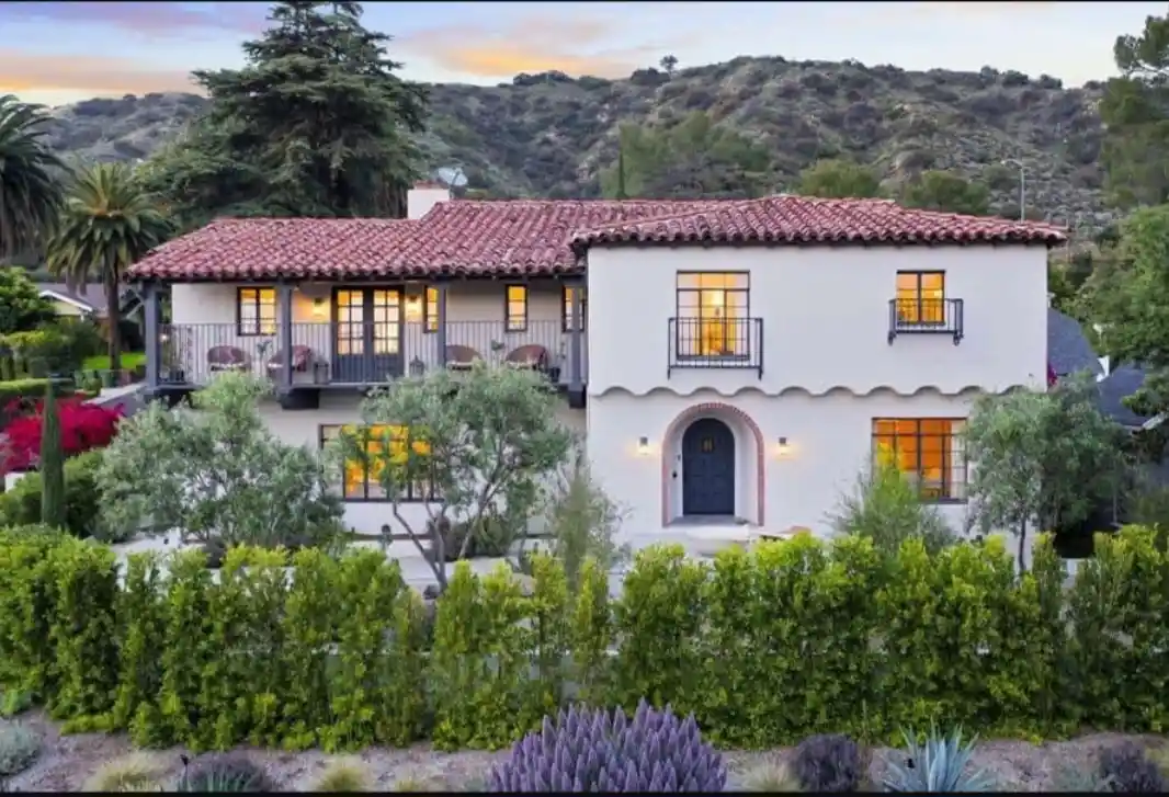 a picturesque two-story white stucco house with red tile roof, arched door, and lit windows, surrounded by lush greenery and…