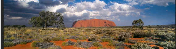 A wide landscape photo of Uluru (Ayers Rock) in Australia under a partly cloudy sky, with sparse desert vegetation and two…