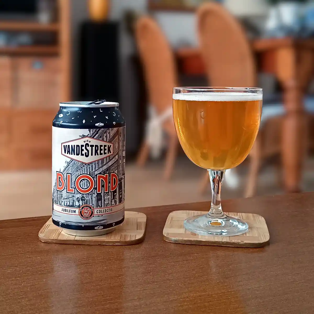 A can of VandeStreek Blond beer sits next to a glass of poured beer on wooden coasters on a wooden table. The can features a…