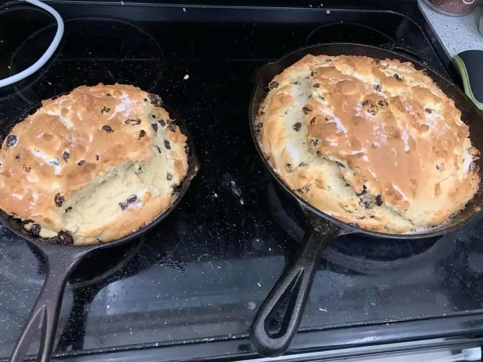 Two cast-iron skillets sit on a stovetop, each containing a golden-brown Irish soda bread dotted with raisins. The bread on…