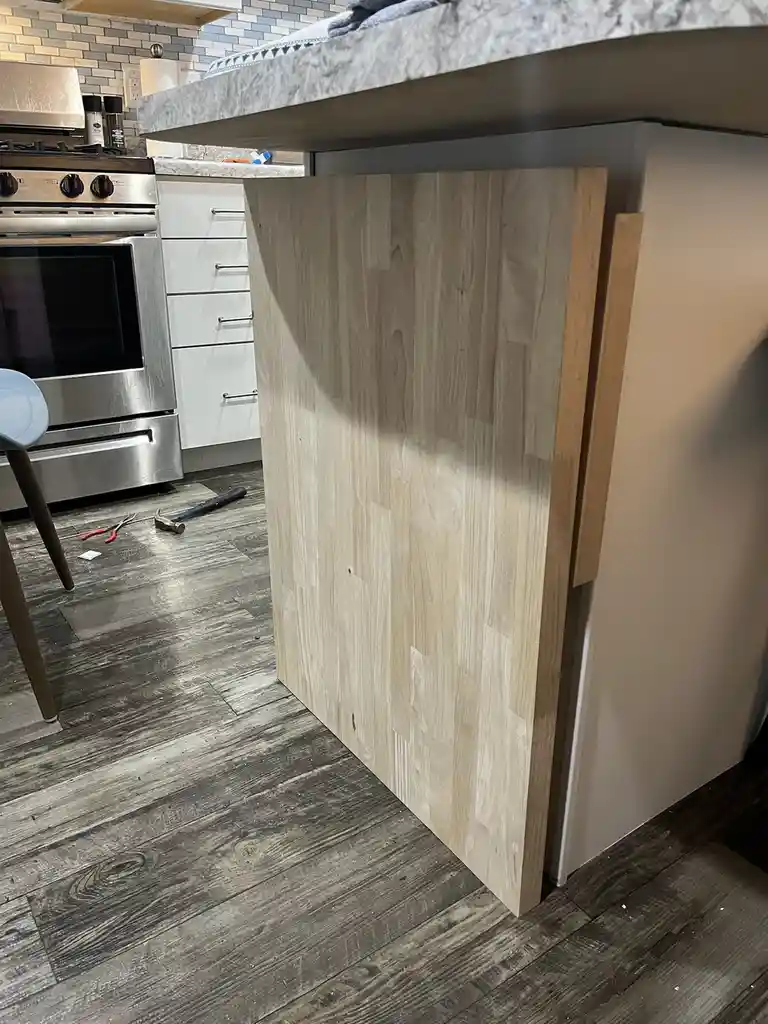 A kitchen scene shows a large, unfinished light-wood panel leaning against a newly installed kitchen island with a marble…