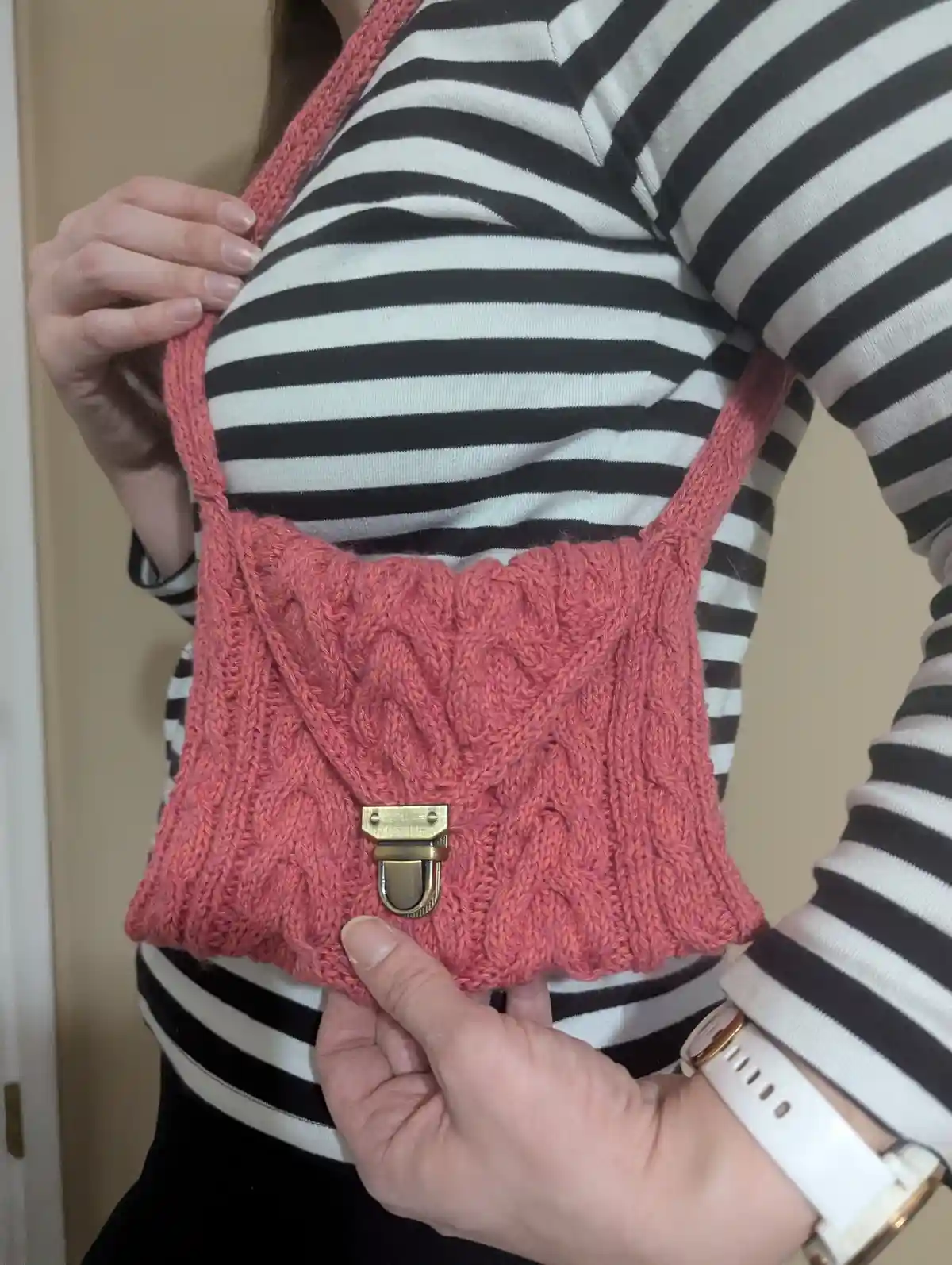 A person wearing a black-and-white striped shirt holds up a handmade coral-colored knitted bag with cable patterns and a…