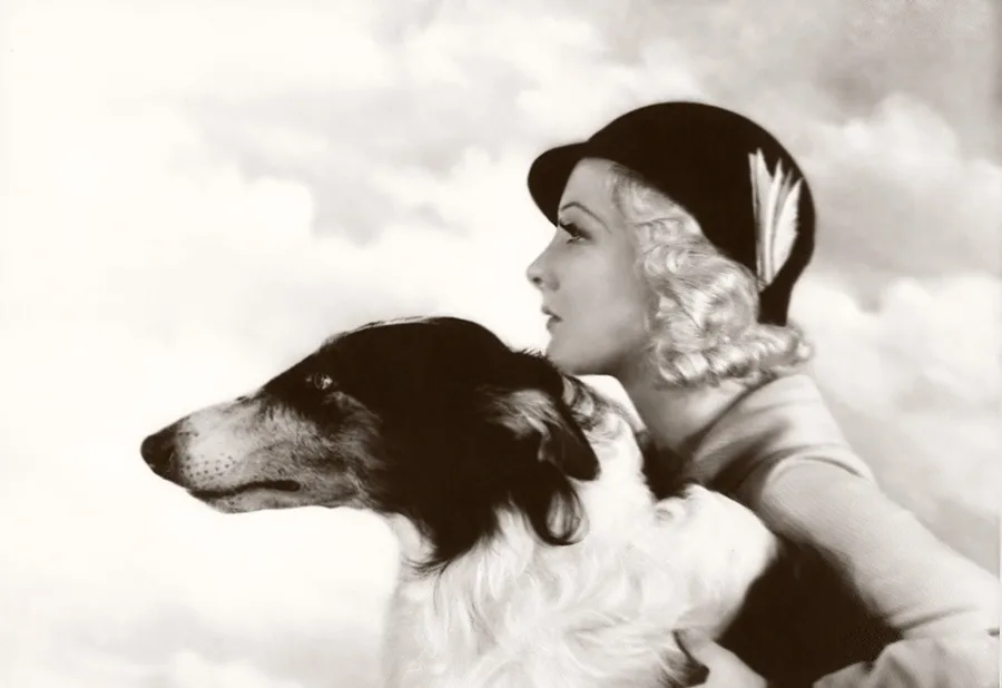 A vintage black-and-white photograph shows a woman with blonde curls and a cloche hat looking pensively to the side, with a…
