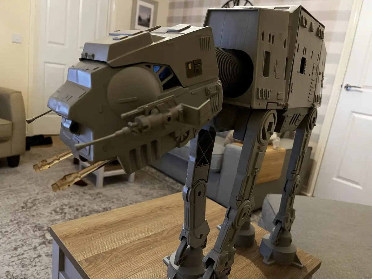 A large, grey plastic AT-AT walker toy from Star Wars is standing on a wooden table in a living room, its front legs and…