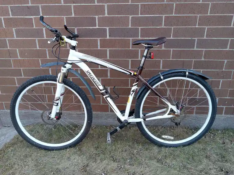A white and brown Specialized Hardrock mountain bike with fenders and disc brakes stands upright on dry grass against a red…