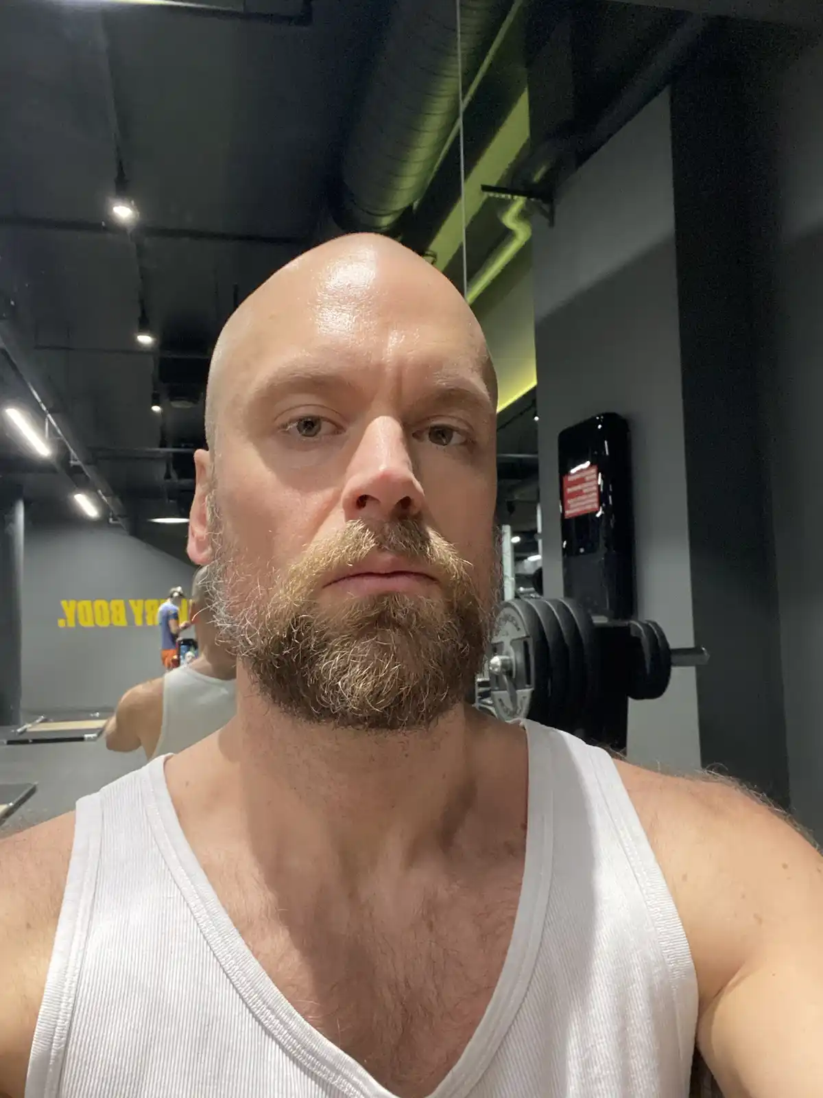 A bald man with a beard, wearing a white tank top, takes a selfie in a gym. Behind him, gym equipment and another person are…