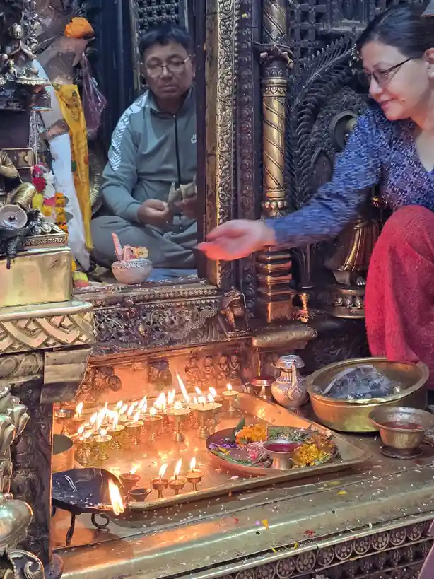 A woman in a blue patterned top and red pants is seen offering something at a temple altar with many lit oil lamps, while a…