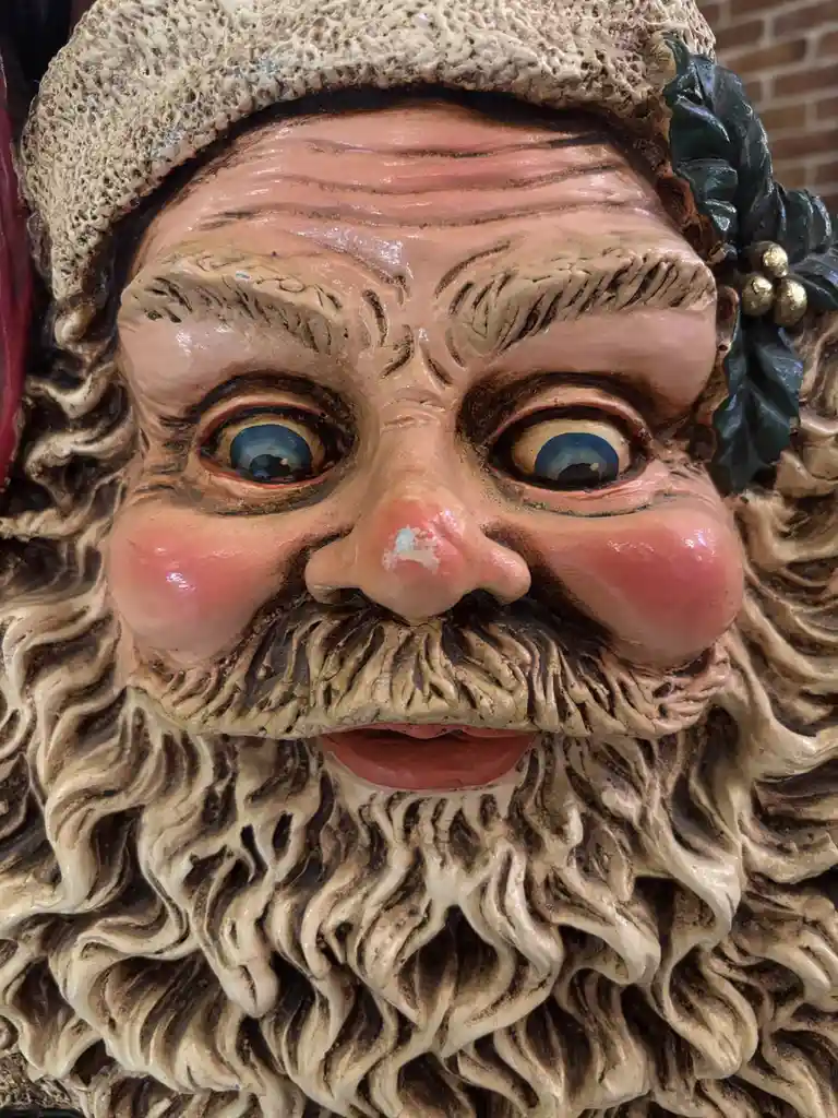 A close-up of a weathered, plastic Santa Claus statue with exaggerated, wide-set blue eyes, a bulbous nose, and a full…