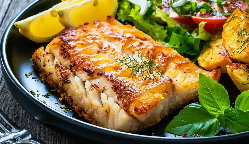 a close-up photo of a golden-brown, crispy-fried fish fillet served on a black plate, accompanied by lemon wedges, a fresh…