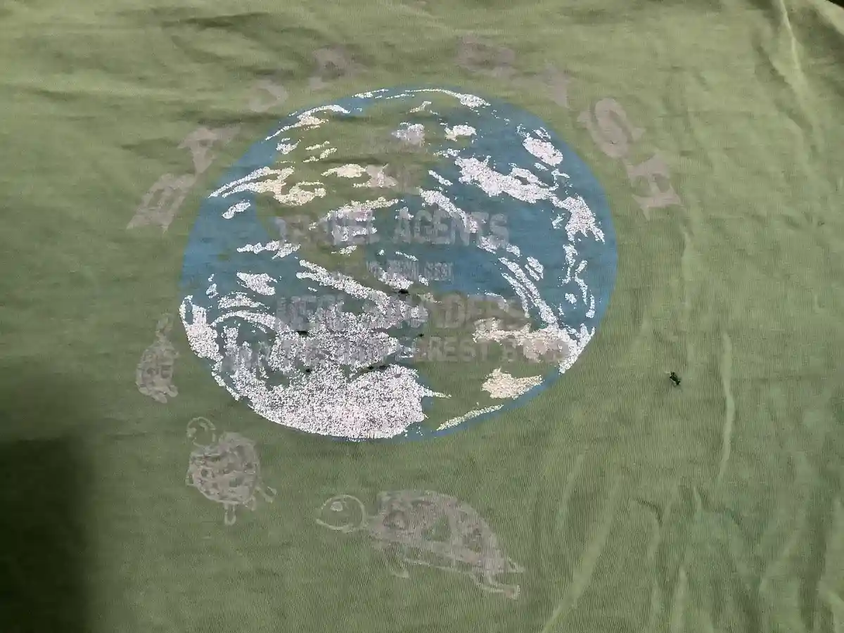 A close-up of a faded green t-shirt with a distressed graphic of a blue and white globe. Around the globe, the words 'BAJA…