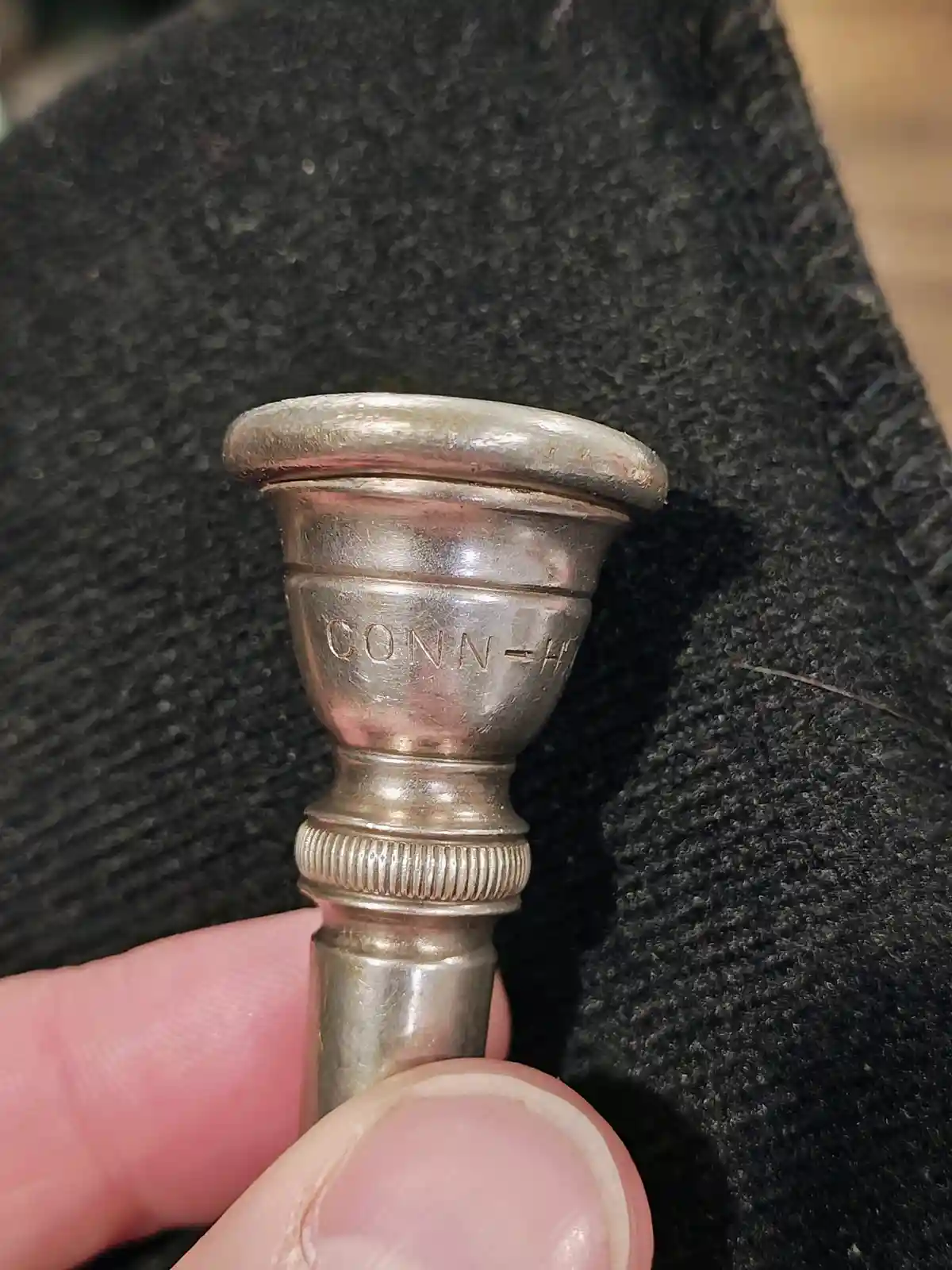 A close-up photo shows a person's fingers holding a tarnished silver-colored brass instrument mouthpiece against a dark gray…